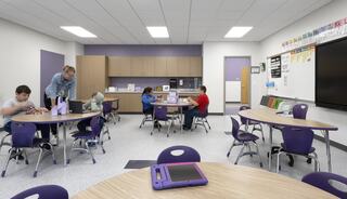 A modern classroom features children engaged in activities at round tables, with a teacher assisting, vibrant walls, and educational tools.