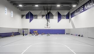 A spacious gymnasium features a basketball hoop, gray flooring, and colorful equipment, with children engaged in activities along the wall.