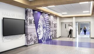 A modern hall features a vibrant mural of a Native American figure alongside historical photos, with sleek design elements and bright lighting.