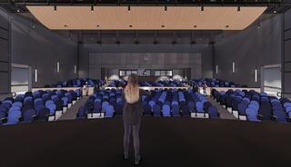 A performer stands on stage, facing a spacious auditorium filled with rows of blue seats, under a modern lighting setup.