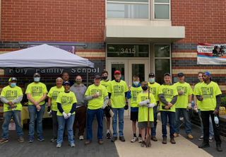 A group of volunteers in bright yellow shirts poses outside a school, smiling and ready for community service with masks and gloves on.