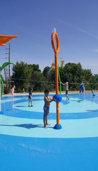 Children play in a colorful splash pad, enjoying water features under a clear blue sky on a sunny day. Excitement and laughter abound.