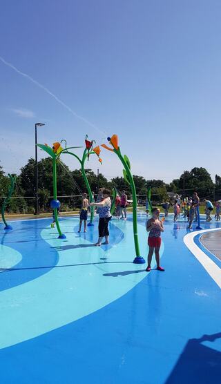 Children play joyfully in a colorful water splash park under a clear blue sky, surrounded by vibrant greenery.