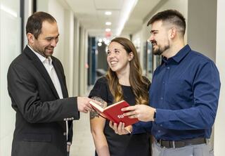 Three professionals engage in conversation while looking at a book, sharing smiles in a well-lit, modern hallway.