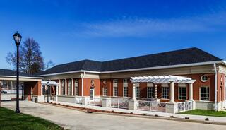A modern brick building with large windows, a white pergola, and landscaped surroundings, set against a clear blue sky.
