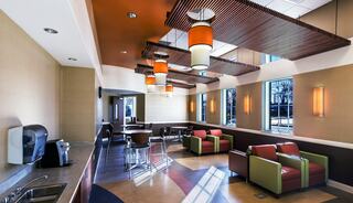 A modern lounge area with colorful seating, decorative lighting, and a coffee machine, featuring large windows that bring in natural light.