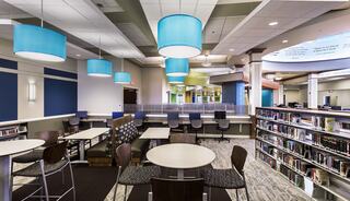 A modern library interior features bright pendant lighting, study tables, comfortable seating, and shelves filled with books.