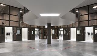 A modern, circular prison corridor with multiple closed doors, open to a central area illuminated by overhead lighting and surrounded by glass walls.