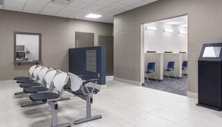 A modern waiting area features white chairs, a partitioned section with blue seating, and a sleek kiosk, all in neutral tones.