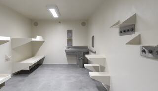 A sparse, sterile room featuring white walls, concrete flooring, shelving, a sink, and a trash can, designed for minimal comfort.