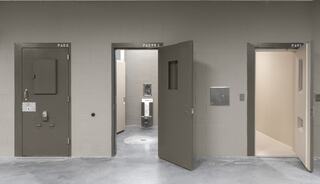 A stark, institutional hallway features three heavy doors labeled PAD1, PAD2, and PADR1. One door stands open, revealing a bleak cell interior.