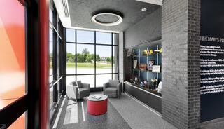 A modern, bright lobby features two gray chairs, a red table, and a showcase filled with items, illuminated by large windows.