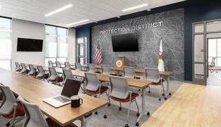 A modern conference room with wooden tables, ergonomic chairs, an American flag, and a wall featuring the Godfrey Fire Protection District logo.