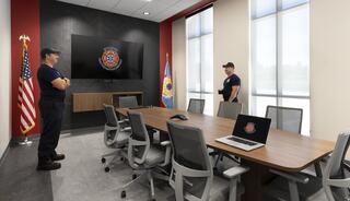A modern conference room features two firefighters, a large screen, a flag, and a state flag, emphasizing collaboration and professionalism.
