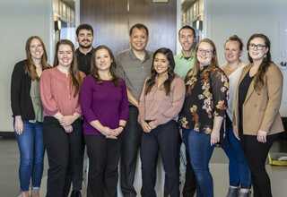 A group of ten individuals, smiling and posing together in a bright office setting, showcasing a diverse range of clothing styles.