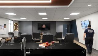 A modern lounge with comfortable seating, a large TV, and a sleek kitchen counter. A firefighter walks past, adding a communal touch.