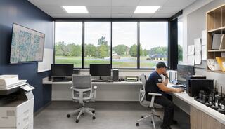 A modern office features two desks with computers, a printer, and a large window showcasing greenery outside, while a person works attentively.