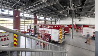 A modern fire station interior with multiple fire trucks parked, large windows for natural light, and an organized, clean workspace.