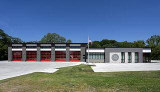 A modern fire station features large red garage doors, a brick facade, and a flag, set against a backdrop of green trees and blue skies.