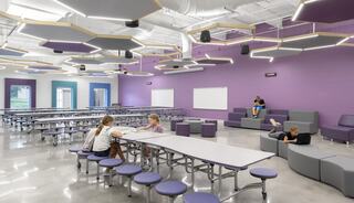 A modern, spacious cafeteria features purple walls, flexible seating, and bright lights, with students interacting and relaxing in the space.