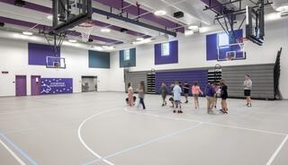 A bright gymnasium features a group of children playing, with basketball hoops and bleachers in the background, creating an active atmosphere.