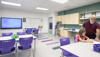 A brightly lit classroom features purple chairs and tables. A teacher assists a student, while another child works at a sink.
