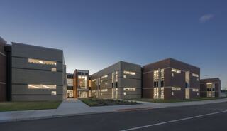 A modern, multi-story building features large windows and a sleek design, set against a twilight sky and accompanied by landscaped greenery.