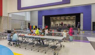 A bright school cafeteria with children sitting at tables, some eating, while others play. A stage features a group performing in the background.