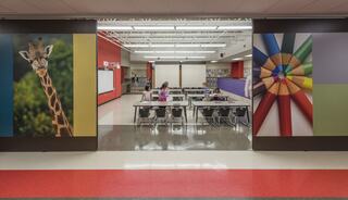 A vibrant hallway features large images of a giraffe and colored pencils, leading to a classroom with empty desks and colorful walls.