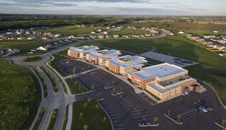 A modern building sits amidst lush green fields and residential areas, featuring ample parking and well-maintained surroundings.