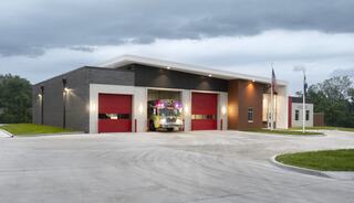 A modern fire station featuring large red doors, a fire truck in front, and flags outside, set against a cloudy sky.