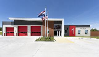 A modern fire station features red doors, large windows, and flagpoles displaying the U.S. and state flags, with a prominent number "1."