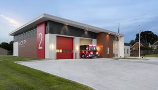 A modern fire station with a vibrant red door, featuring a fire truck ready for action and flags outside, set against a twilight sky.