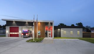 A modern fire station at dusk, featuring a red garage door, flagpoles, and a fire truck, surrounded by a manicured lawn.