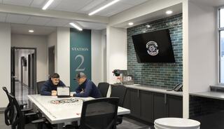 Two individuals sit at a table in a modern room with a green tiled wall, a digital screen, and a coffee station, labeled "Station 2."