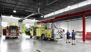 A spacious fire station features two fire trucks, one yellow and one red, with firefighters discussing equipment in a well-lit area.