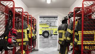 Firefighter gear hangs on red racks, with a glimpse of a yellow fire truck visible through a doorway. The space is organized and ready for use.