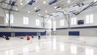 A spacious gymnasium with high ceilings, basketball hoops, and groups of people seated on the court, focusing on a speaker in front.
