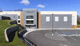 A modern gymnasium with a sleek design, featuring large windows and a landscaped entry, surrounded by a spacious parking lot under a blue sky.