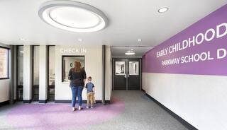 A woman and a child approach a check-in area in a brightly lit school hallway, with purple walls displaying "Early Childhood Parkway School District."