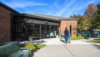 A person walks alongside a child towards a building entrance, surrounded by greenery, flowers, and a clear blue sky.
