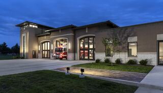 A modern fire station features large garage doors, a well-lit exterior, and a fire truck positioned ready for emergencies at dusk.