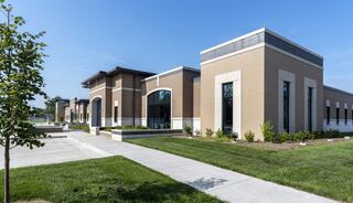 A modern building features a mix of earthy tones, large windows, and landscaped walkways surrounded by lush green grass under a clear sky.