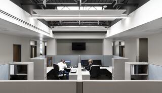 A modern office features minimalistic cubicles, large windows, and two employees working at their desks under bright overhead lighting.