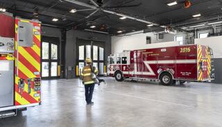 A firefighter in uniform walks through a spacious, modern fire station with fire trucks parked inside and large doors for access.