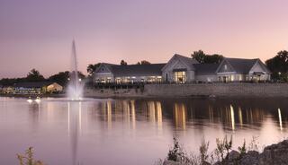 A serene lake reflects a charming building at twilight, with a gentle fountain adding tranquility to the peaceful scene. Trees frame the view.