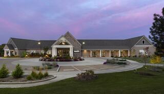 A modern building with a stone facade, surrounded by landscaped gardens and colorful flowers, set against a twilight sky.