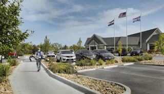 A person walks along a path carrying an object, with several parked cars and flags flying near a modern building in a landscaped area.
