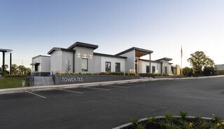 A modern building features a sleek design with large windows, surrounded by greenery and a spacious parking area. An American flag stands nearby.