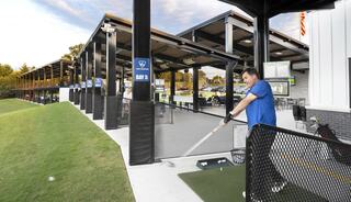 A man swings a golf club at a driving range, with several batting bays and a grassy area, under a clear sky.